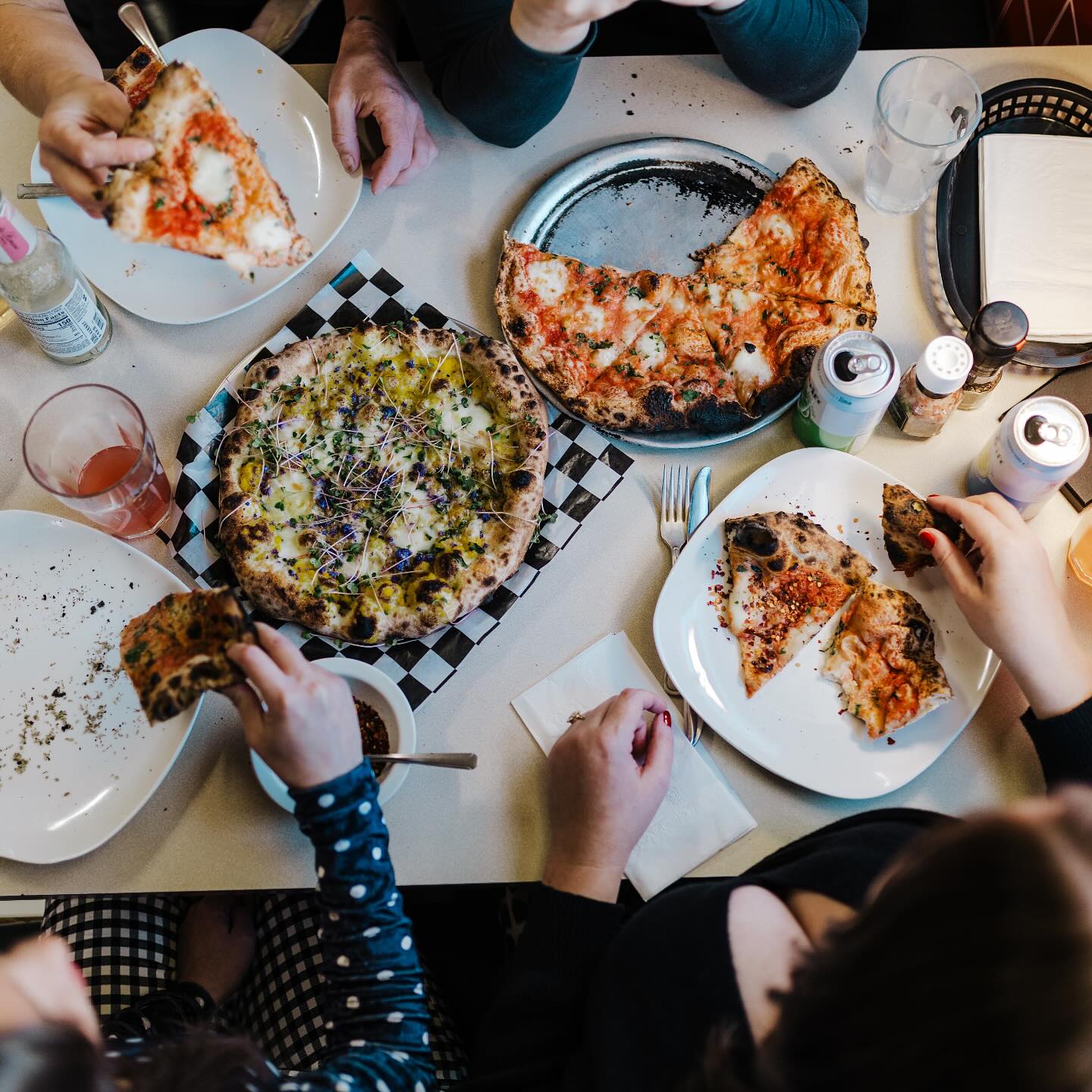 Overhead table scene — multiple pies, slices in hand, brewery cans, checkered placemats